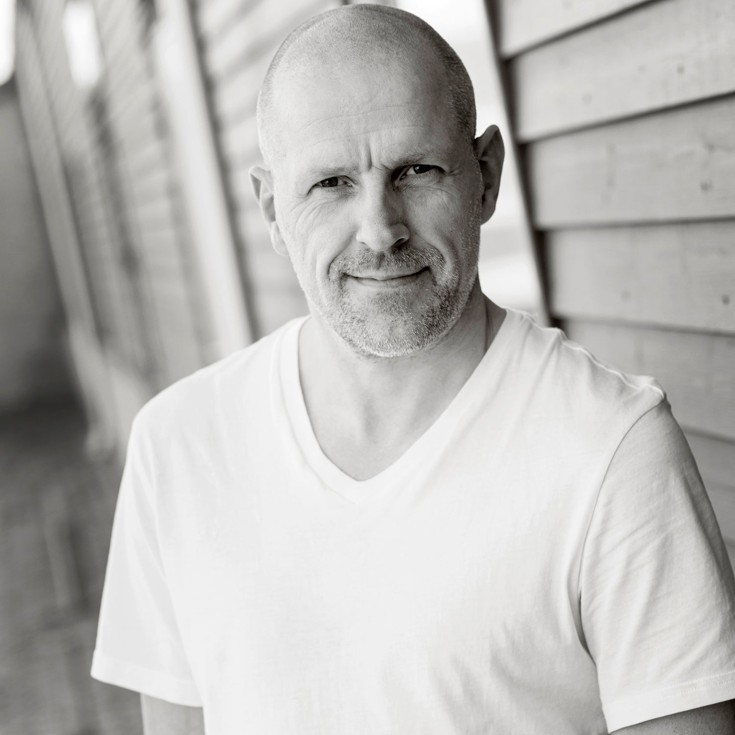 Mann mit Glatze in weißem T-Shirt schaut in die Kamera, neben Holzverkleidung stehend.