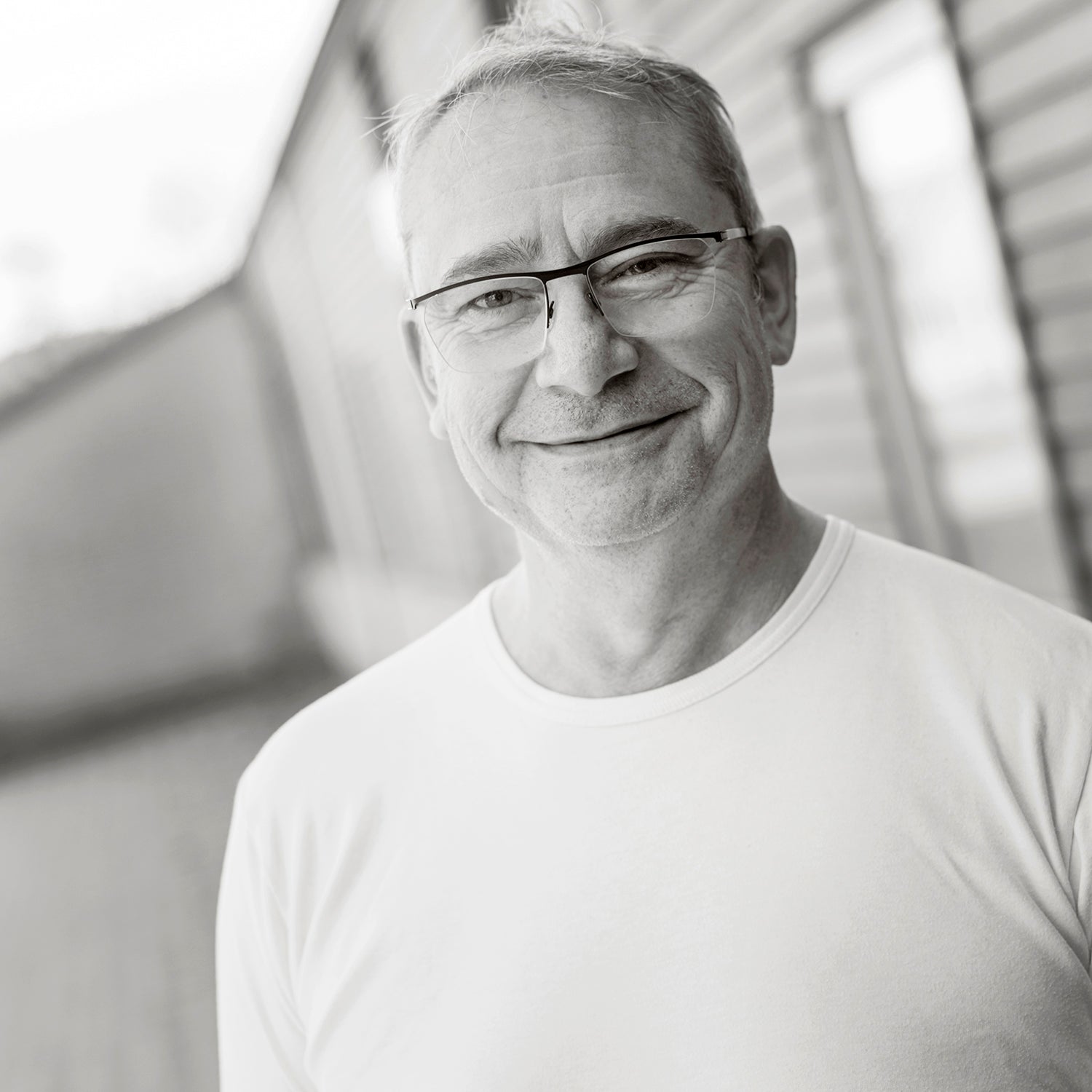 Mann mit Brille und weißem T-Shirt lächelt in die Kamera, neben Holzverkleidung stehend.