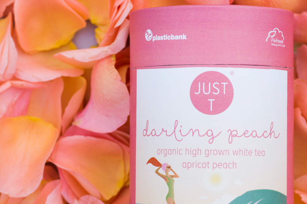 Rosafarbene Teeverpackung mit der Aufschrift "JUST T darling peach" vor rosa Blütenblättern.