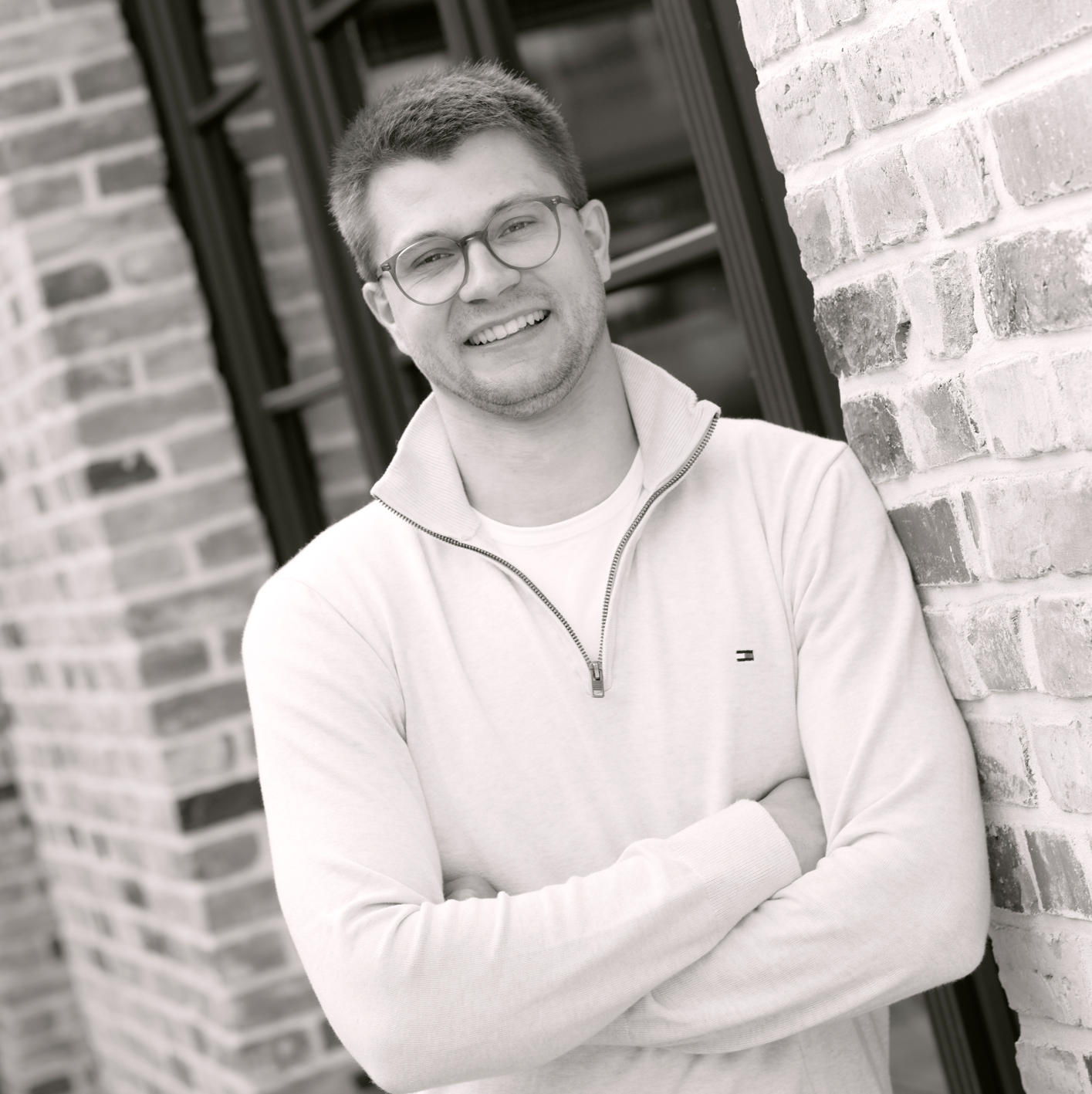 Mann mit Brille und hellem Pullover lehnt lächelnd an einer Backsteinmauer.