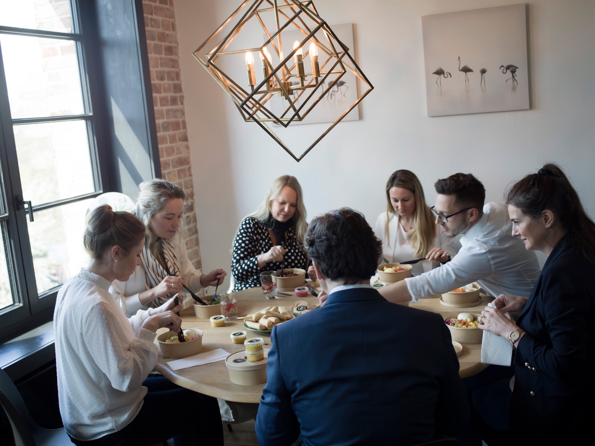 Gruppe von Menschen sitzt um einen Tisch, isst und unterhält sich unter moderner Lampe.