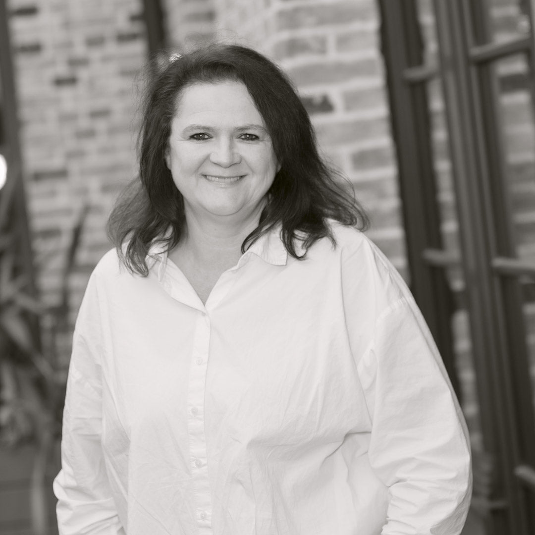 Frau mit dunklem, schulterlangem Haar und hellem Oberteil lächelt vor Backsteinwand.