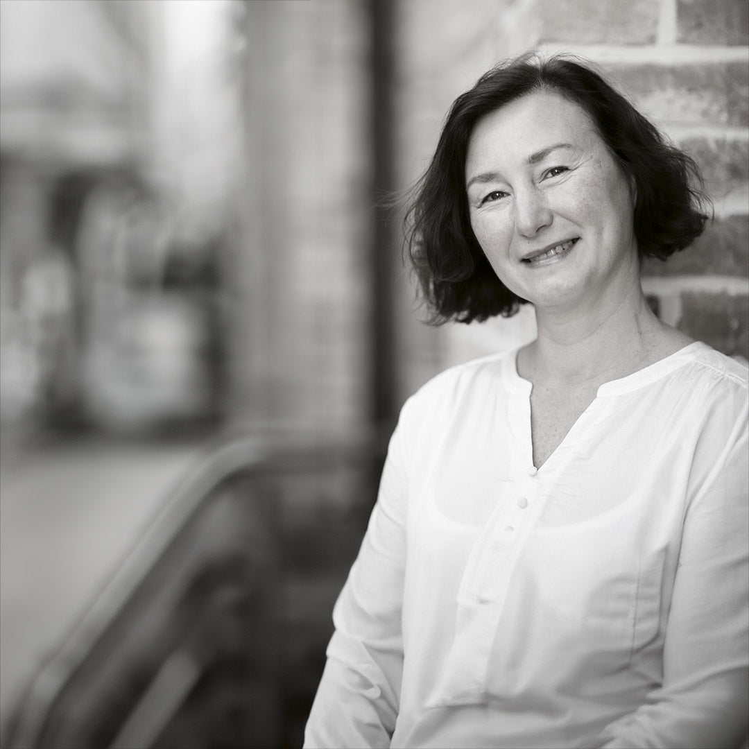 Frau mit schulterlangem dunklem Haar trägt ein weißes Oberteil und lehnt an einer Backsteinwand.