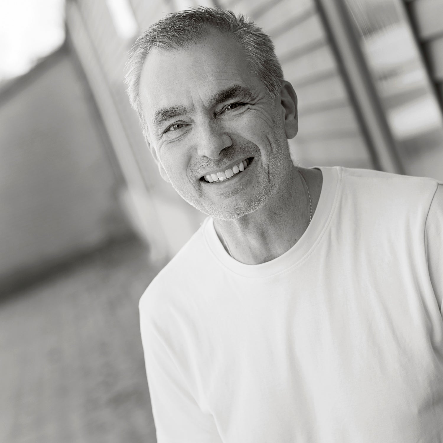 Mann in weißem T-Shirt lächelt in die Kamera, steht vor unscharfem Hintergrund.
