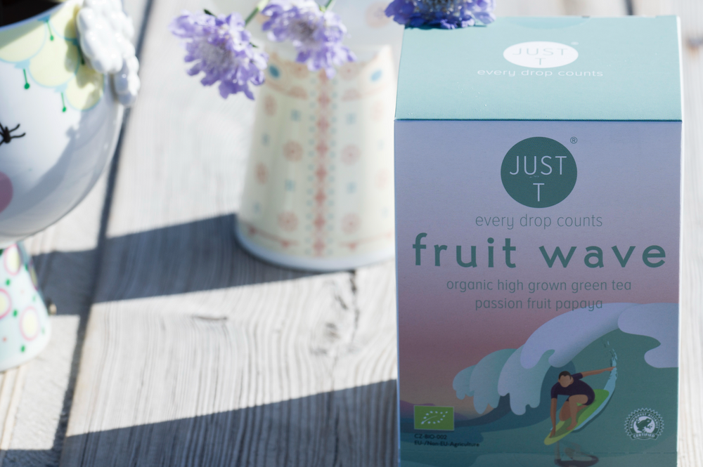 Teeverpackung mit dem Text "Just T fruit wave organic high grown green tea" auf Holztisch mit Blumen.